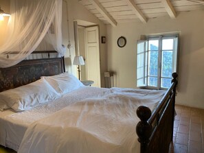 Un dormitorio con una cama grande, dosel, lámpara de noche, ventana con vistas y techo de madera.