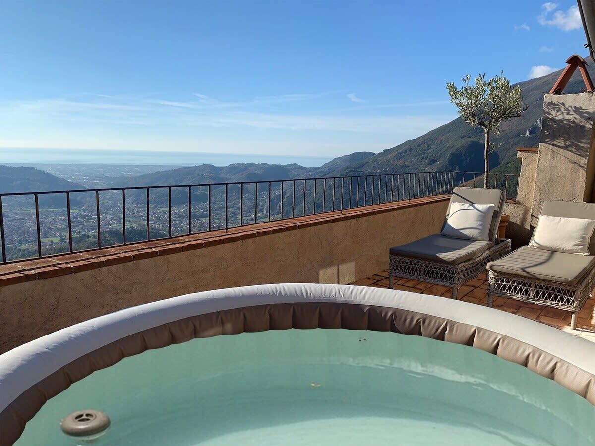 A hot tub with a view of a mountainous landscape, a railing, and two wicker lounge chairs with cushions.