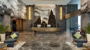 A lobby with a reception desk, two sofas with cushions, a large potted plant, and a wall with a mountainous mural. - The Manohara Hotel Yogyakarta (Depok)