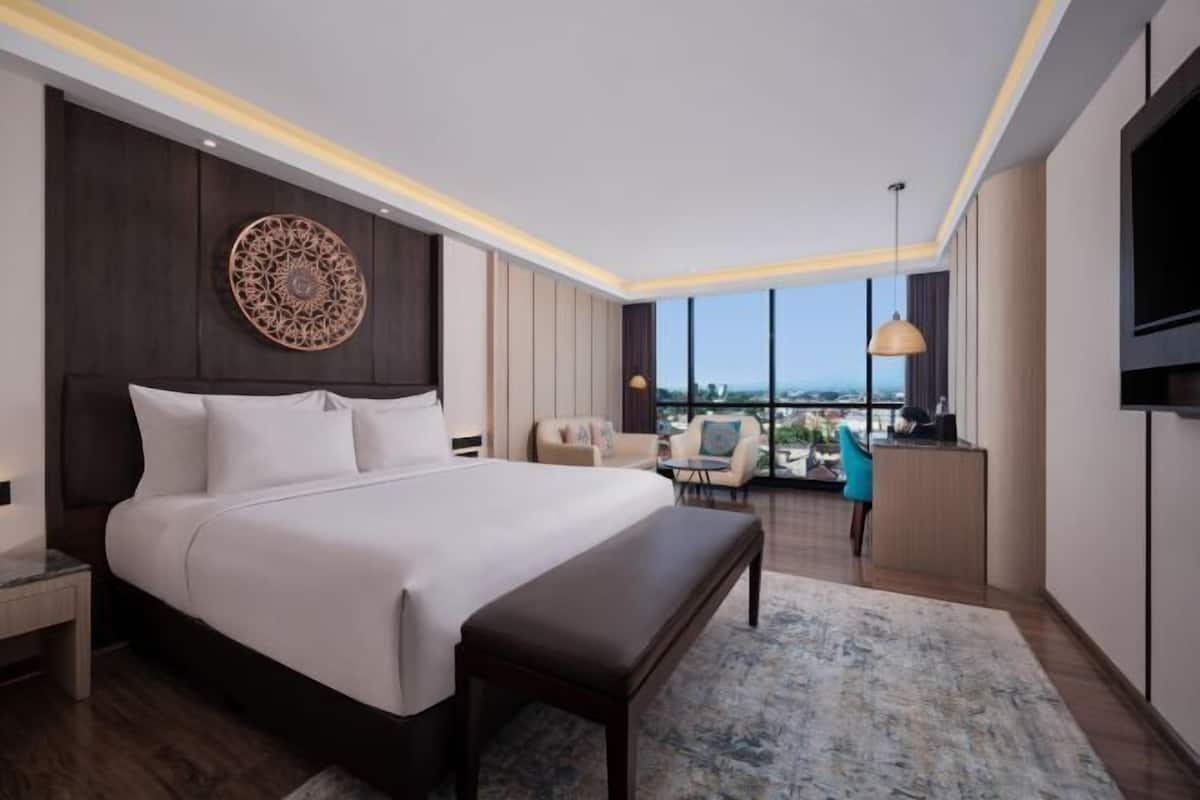 A modern hotel room with a large bed, a desk, a chair, and a view of the city.