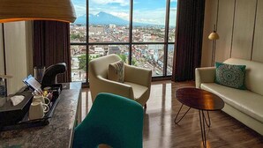 A modern living room with a city view, a sofa, a chair, a coffee table, and a desk with a lamp. - The Manohara Hotel Yogyakarta (Depok)