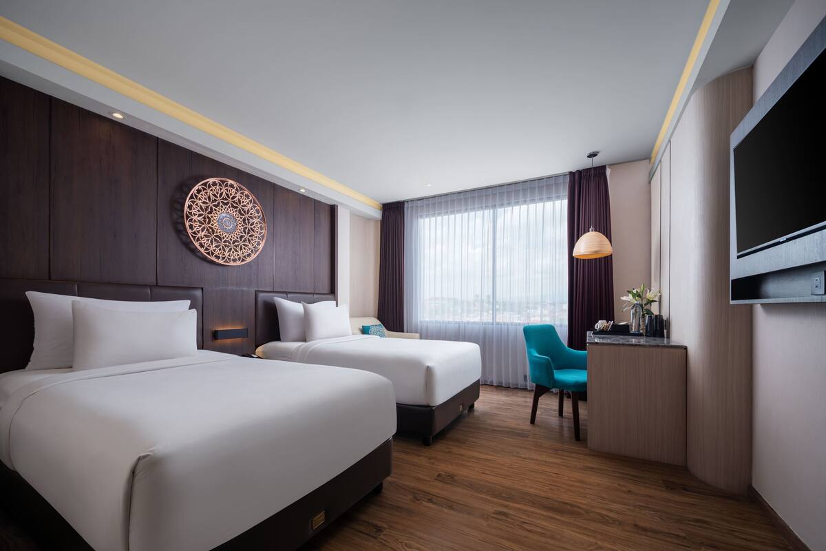 A hotel room with two beds, a desk with a TV, and a large window with curtains.