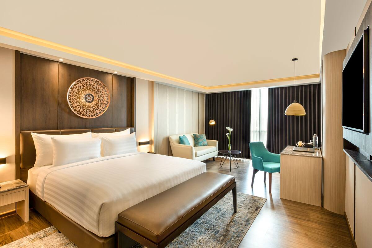 A modern hotel room with a large bed, a seating area with a sofa and armchairs, a small desk, and a television.