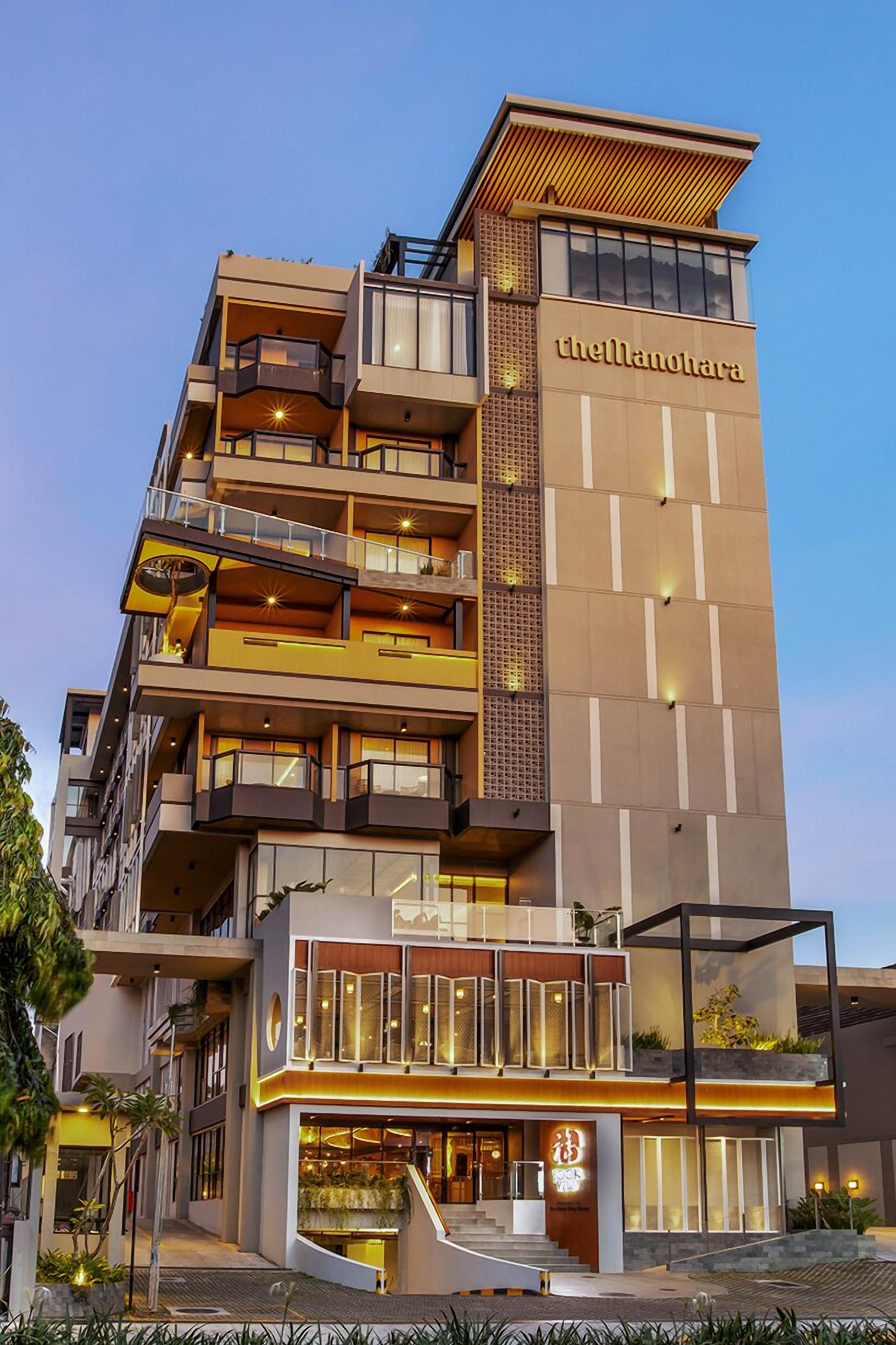A multi-story hotel with a prominent sign reading "theManohara." It features balconies, a ground-level entrance, and a modern architectural design.