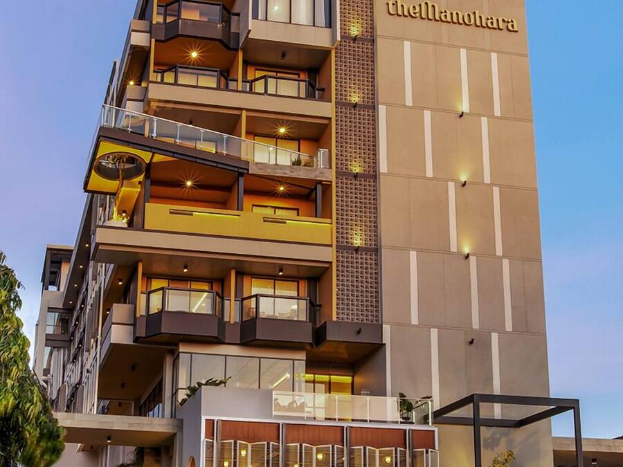 A multi-story hotel with a prominent sign reading "theManohara." It features balconies, a ground-level entrance, and a modern architectural design.
