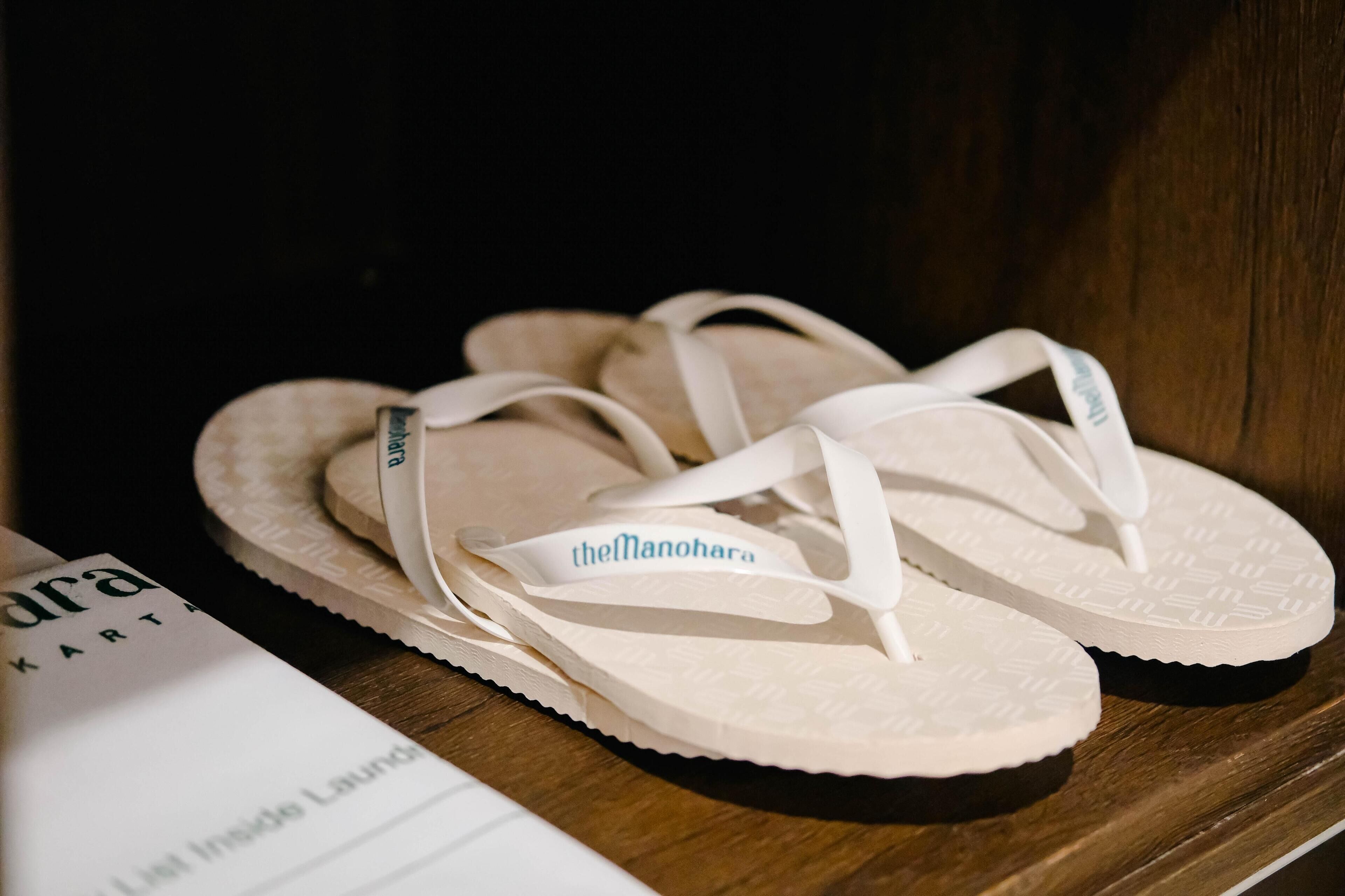 A pair of white flip-flops with a strap labeled "theManohara" on a wooden surface.