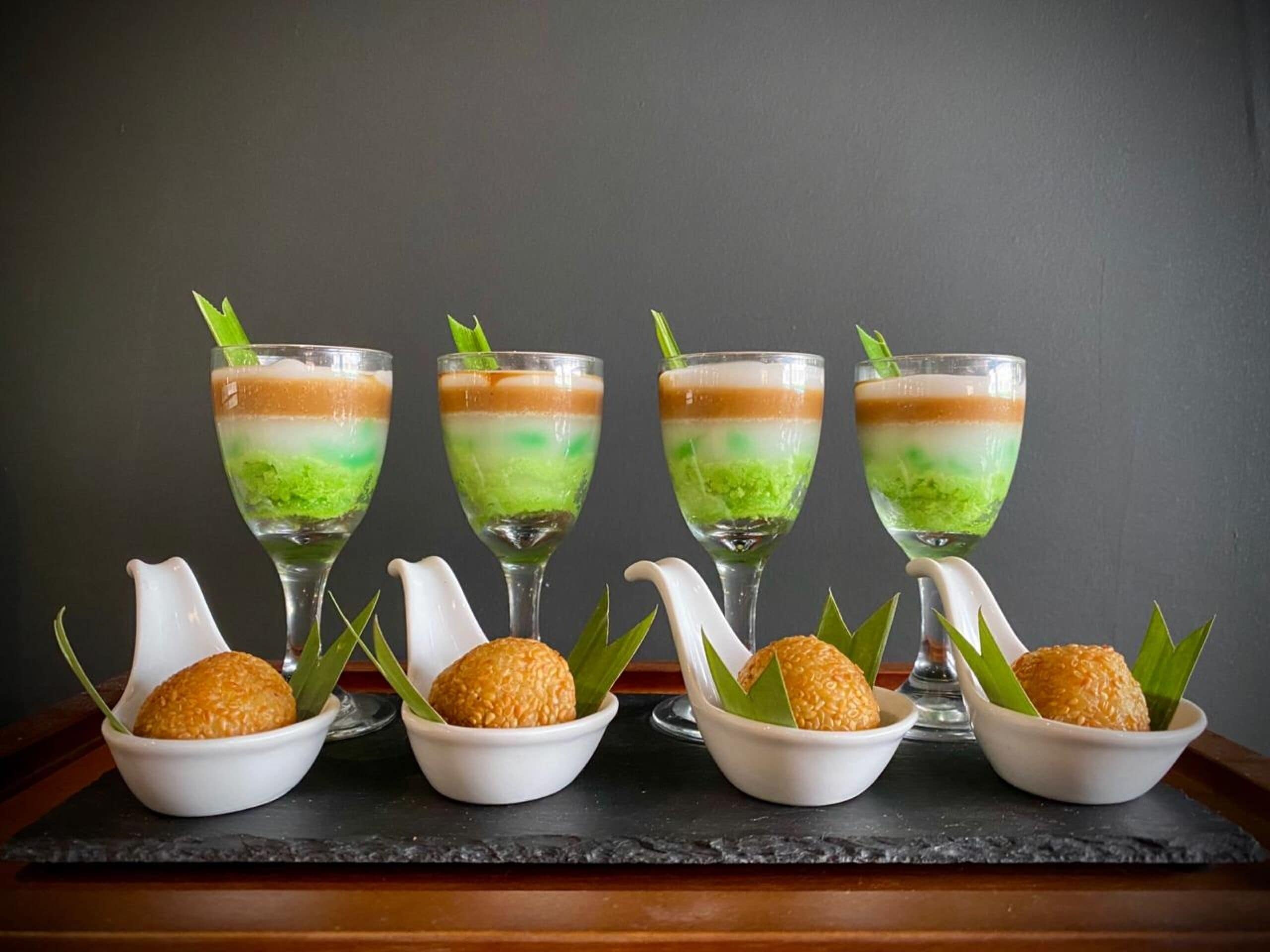 Four glasses of layered dessert with a green topping, each with a white teapot and a bowl of round, golden-brown balls.