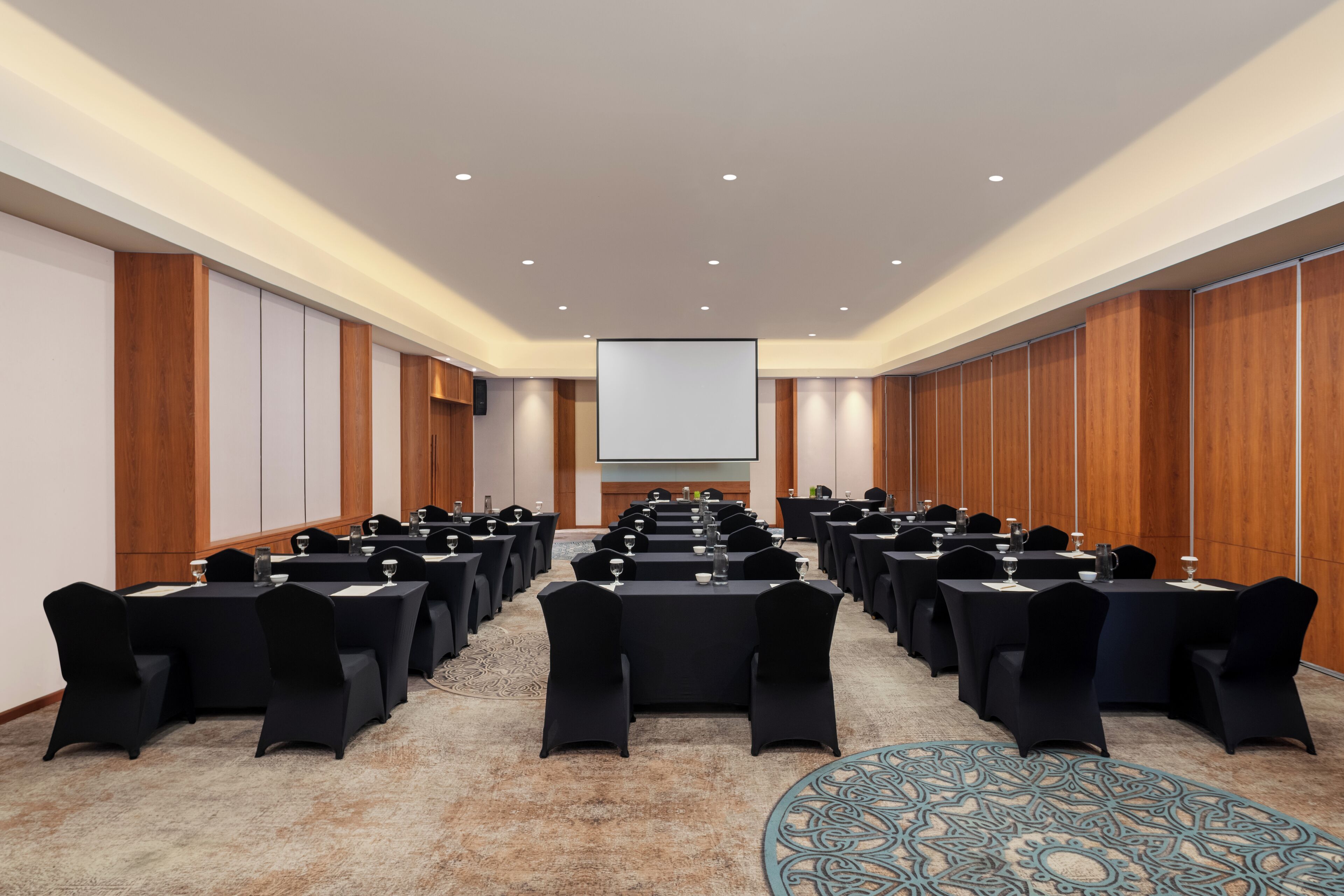 A conference room with rows of tables and chairs, a projector screen, and a carpeted floor.