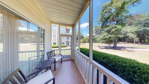 A covered outdoor area with a bench and a table, overlooking a well-maintained lawn and a building with multiple balconies.