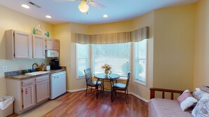 A compact kitchen with wooden cabinets, a microwave, and a coffee maker. There is a dining area with a table and chairs, and a window with blinds.