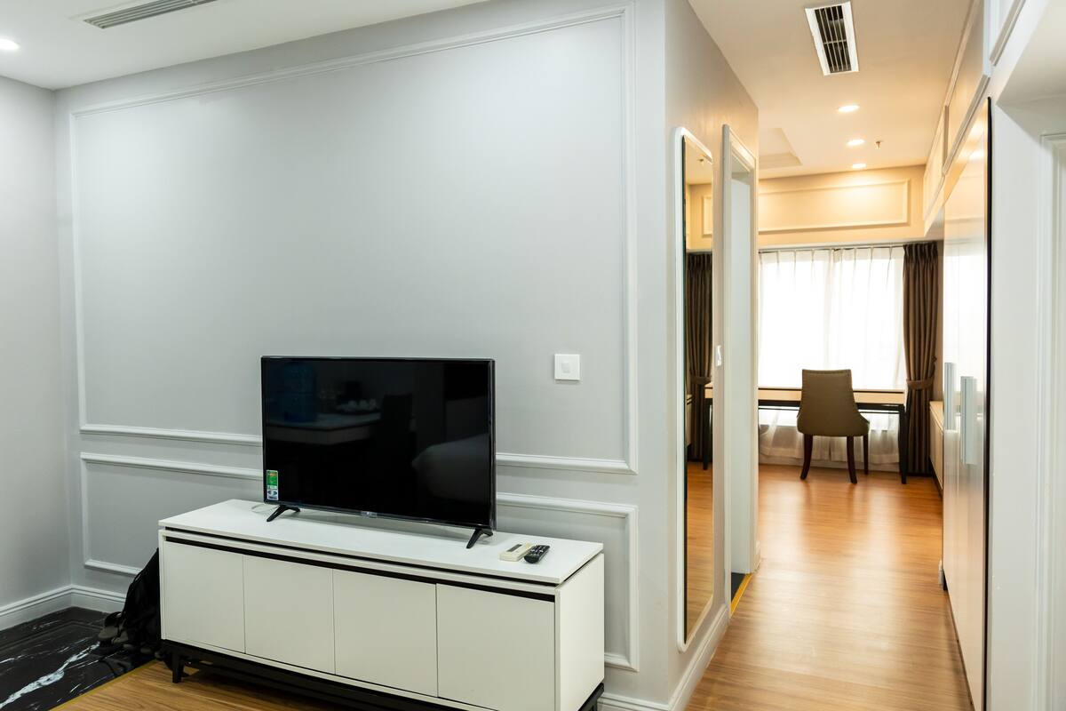Habitación de hotel moderna con televisión de pantalla plana sobre una consola blanca, una pequeña mesa con una silla y vistas al comedor a través de una puerta de vidrio.