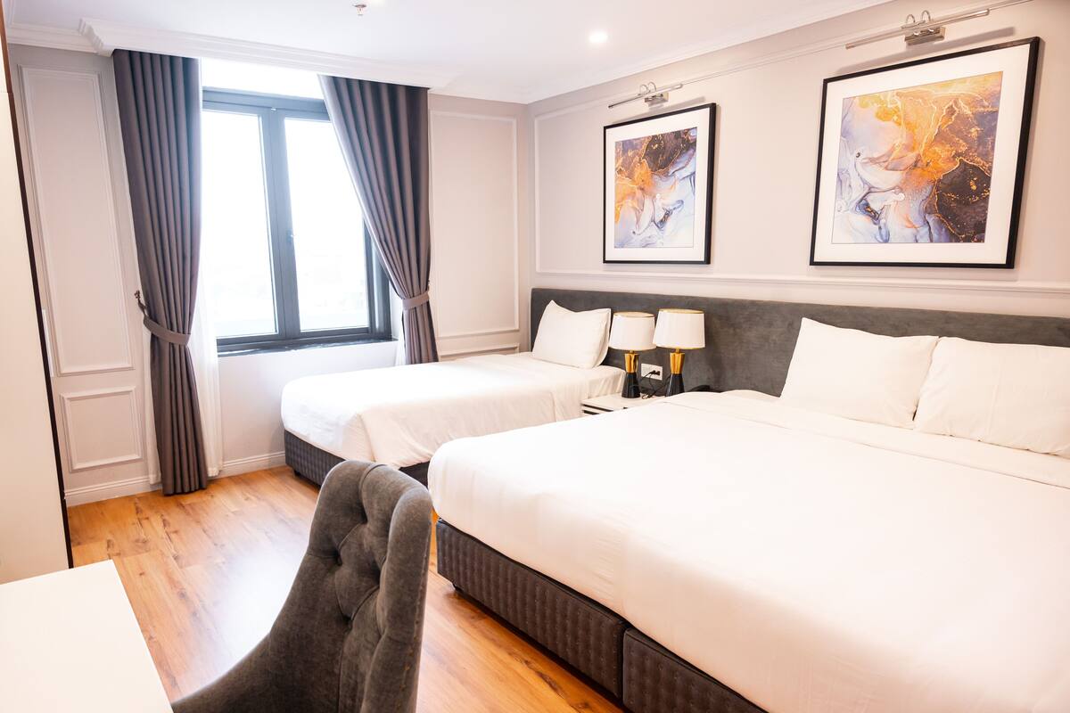 Habitación de hotel con dos camas, un escritorio y una silla. Hay dos cuadros enmarcados en la pared y una ventana con cortinas.