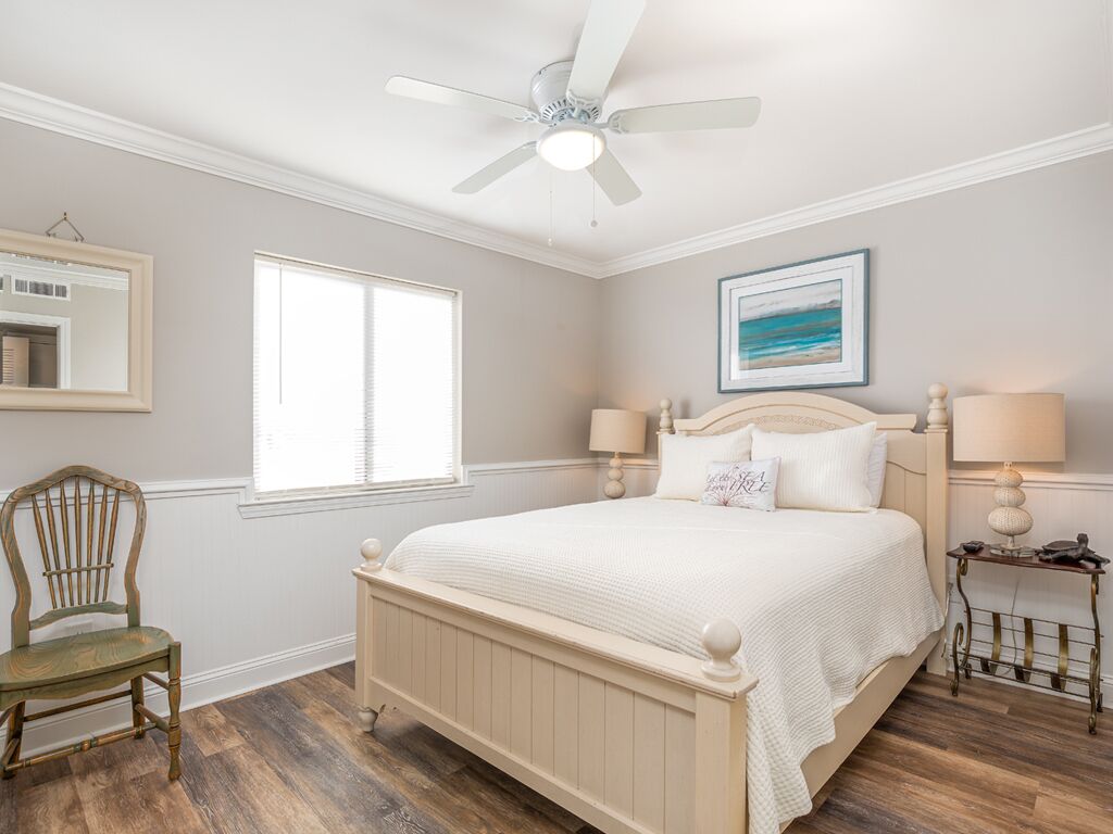 A bedroom with a bed, a chair, a nightstand, a ceiling fan, a window, and a framed picture on the wall.