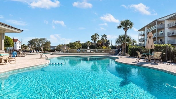 A clear swimming pool with lounge chairs and umbrellas, surrounded by palm trees and a building with balconies. - Lighthouse Point 6C | Pool Views, Beach Access! (Tybee Island)