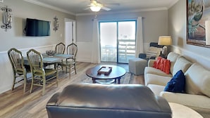 A living room with a sofa, a coffee table, a dining table with chairs, a ceiling fan, and a TV mounted on the wall. - Lighthouse Point 6C | Pool Views, Beach Access! (Tybee Island)
