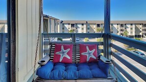 A balcony with a swing seat, two starfish cushions, and a view of a residential area. - Lighthouse Point 6C | Pool Views, Beach Access! (Tybee Island)