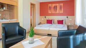 Ein Hotelzimmer mit einem Bett, zwei Nachttischen, einem kleinen Schreibtisch mit Stuhl, einem Sofa, einem Holzschrank mit Küchenutensilien und einer kleinen Pflanze auf dem Schreibtisch.