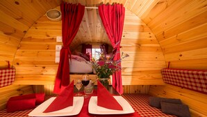 Ein gemütliches Holzhaus mit Essbereich für zwei Personen, ausgestattet mit roten Vorhängen, einer Blumenvase und einer karierten Tischdecke.