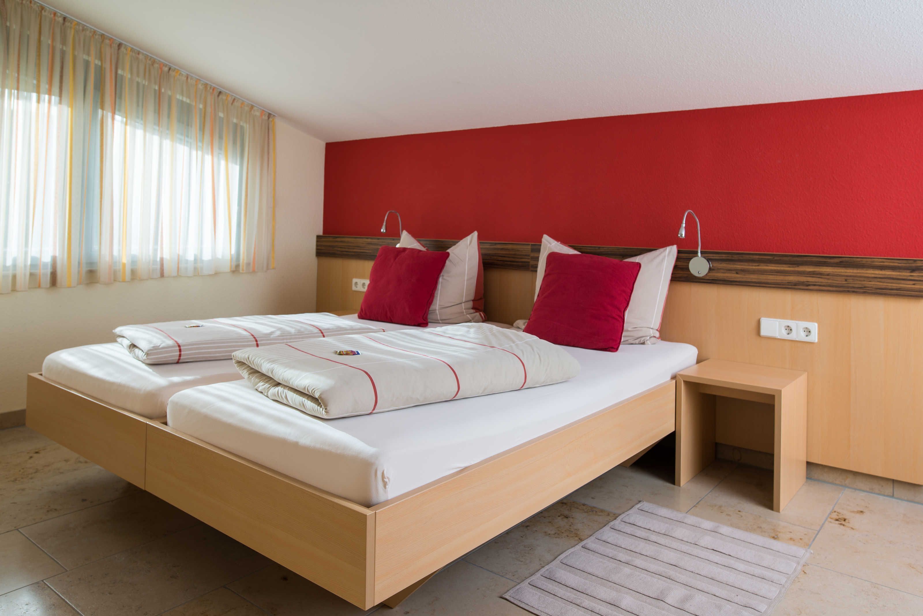 Un lit double avec une literie blanche et des oreillers rouges, une table de chevet en bois et un mur d’accent rouge.
