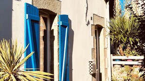 Un bâtiment aux volets bleus, un petit jardin avec des plantes et un ciel bleu dégagé.