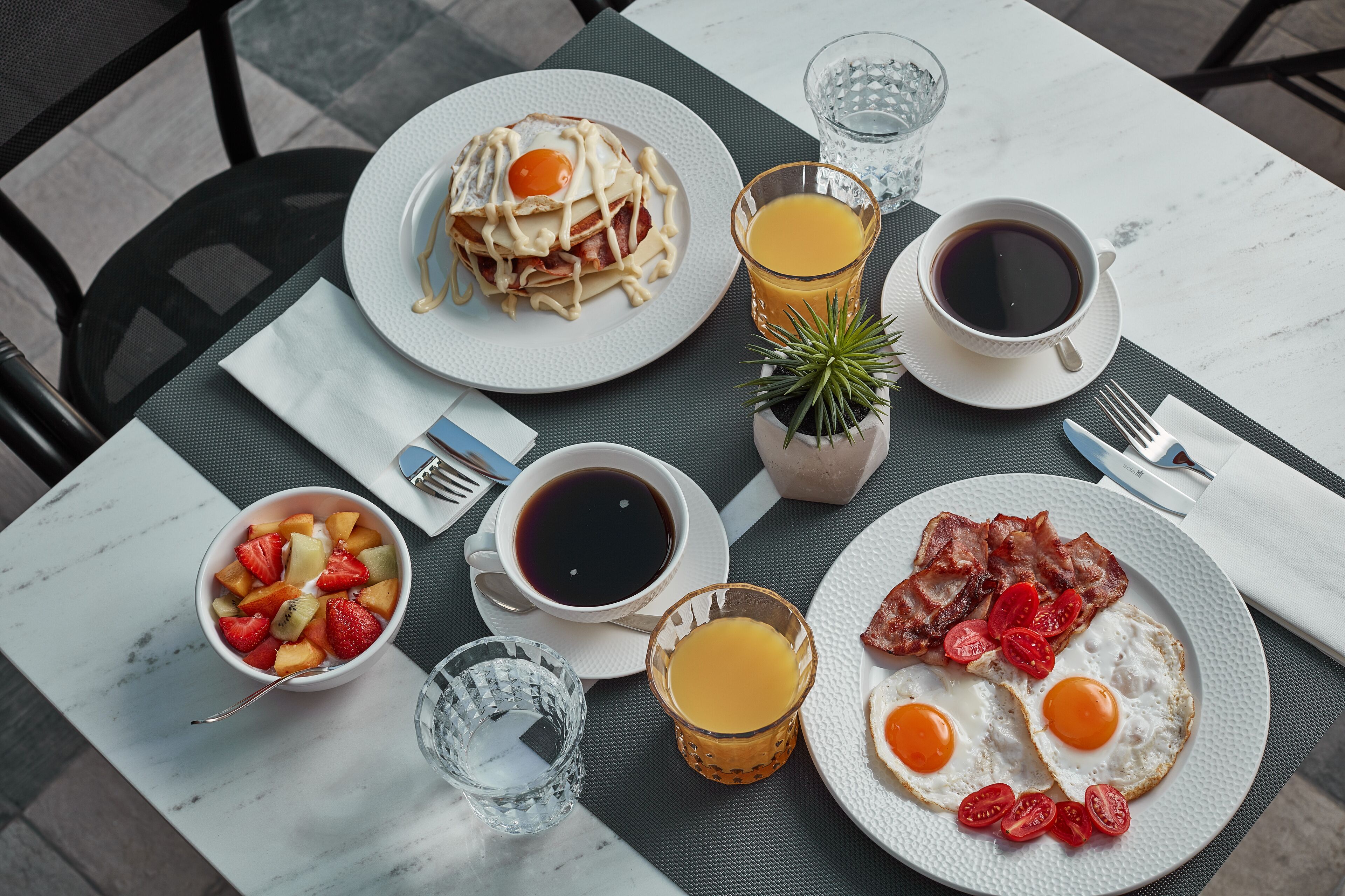 A breakfast table with plates of eggs, bacon, pancakes, and fruit, along with cups of coffee and juice.
