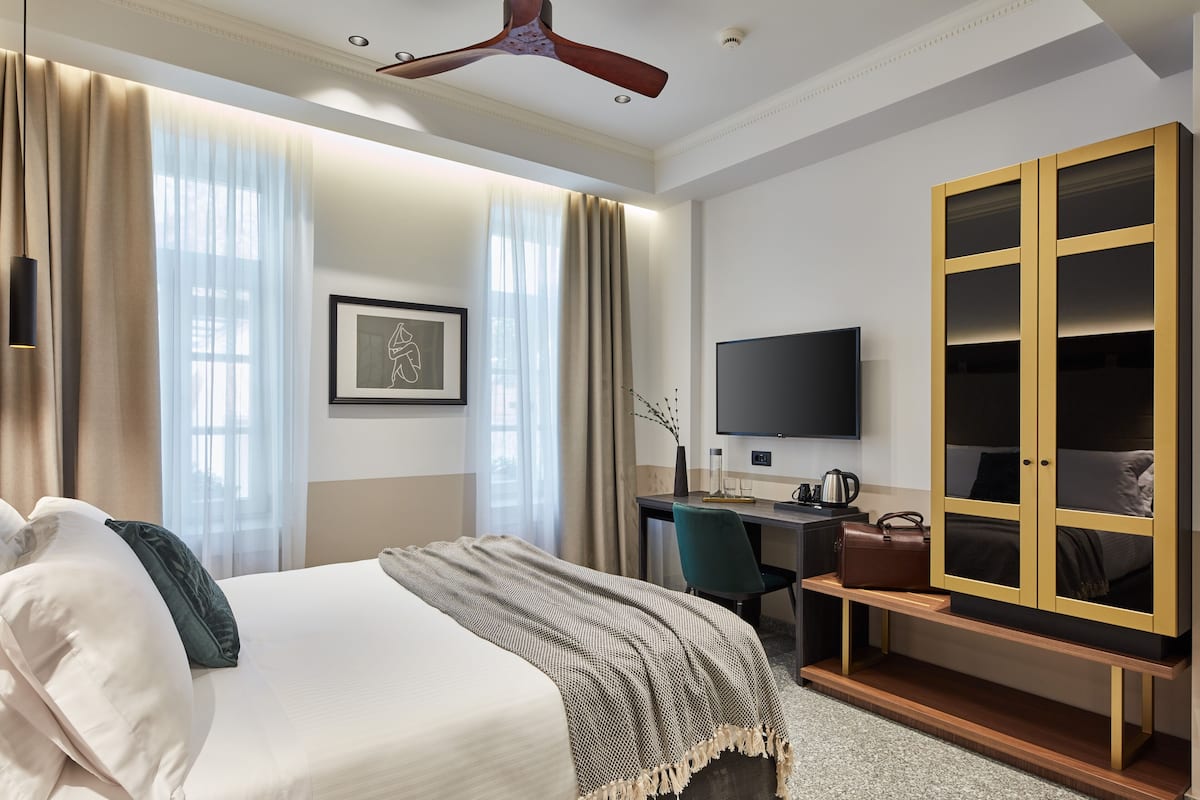 Une chambre d’hôtel comprenant un lit, un bureau avec un ordinateur, une télévision, une armoire et une fenêtre avec des rideaux.