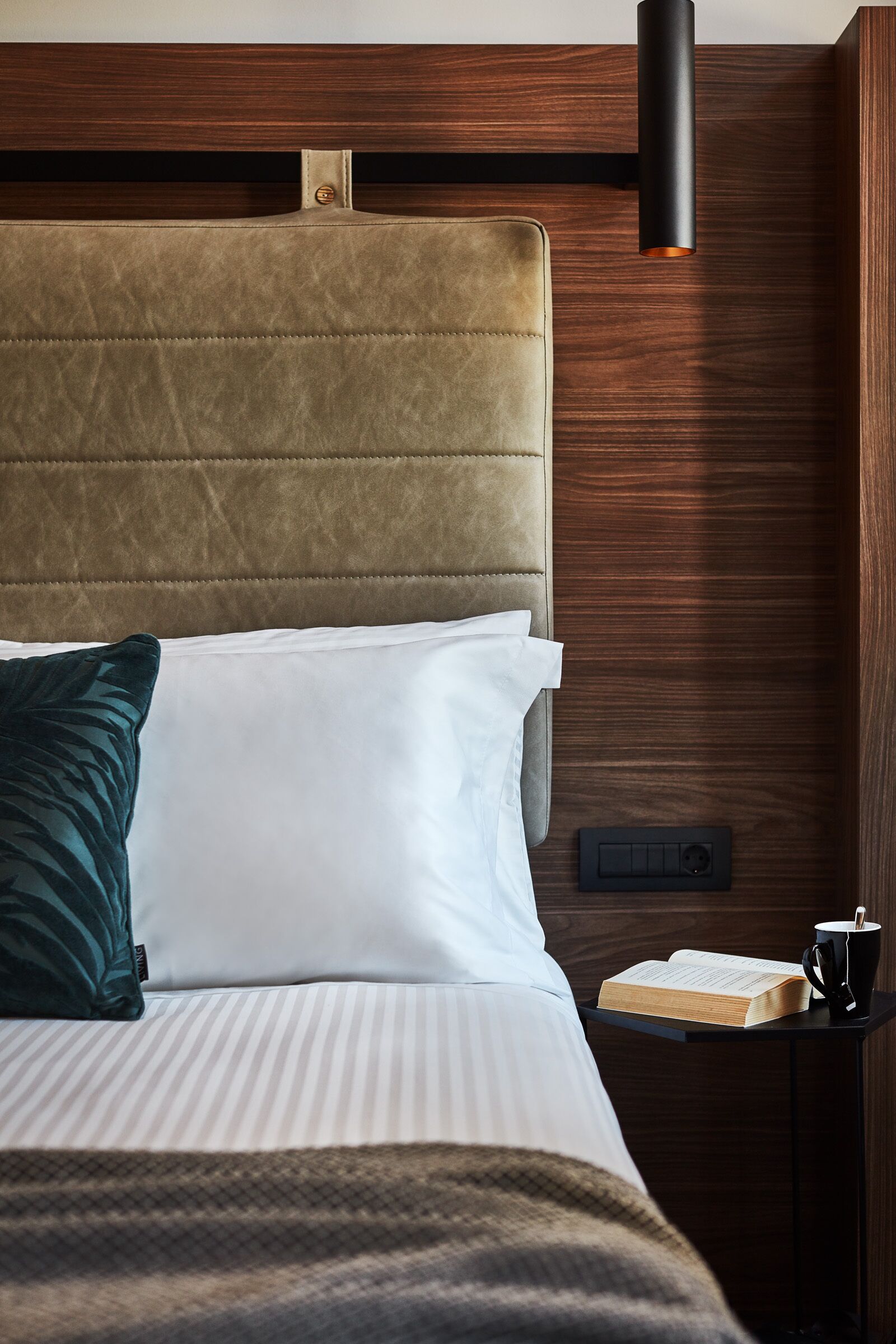 A neatly made bed with a headboard, a bedside table with a book and a mug, and a wall-mounted light fixture.