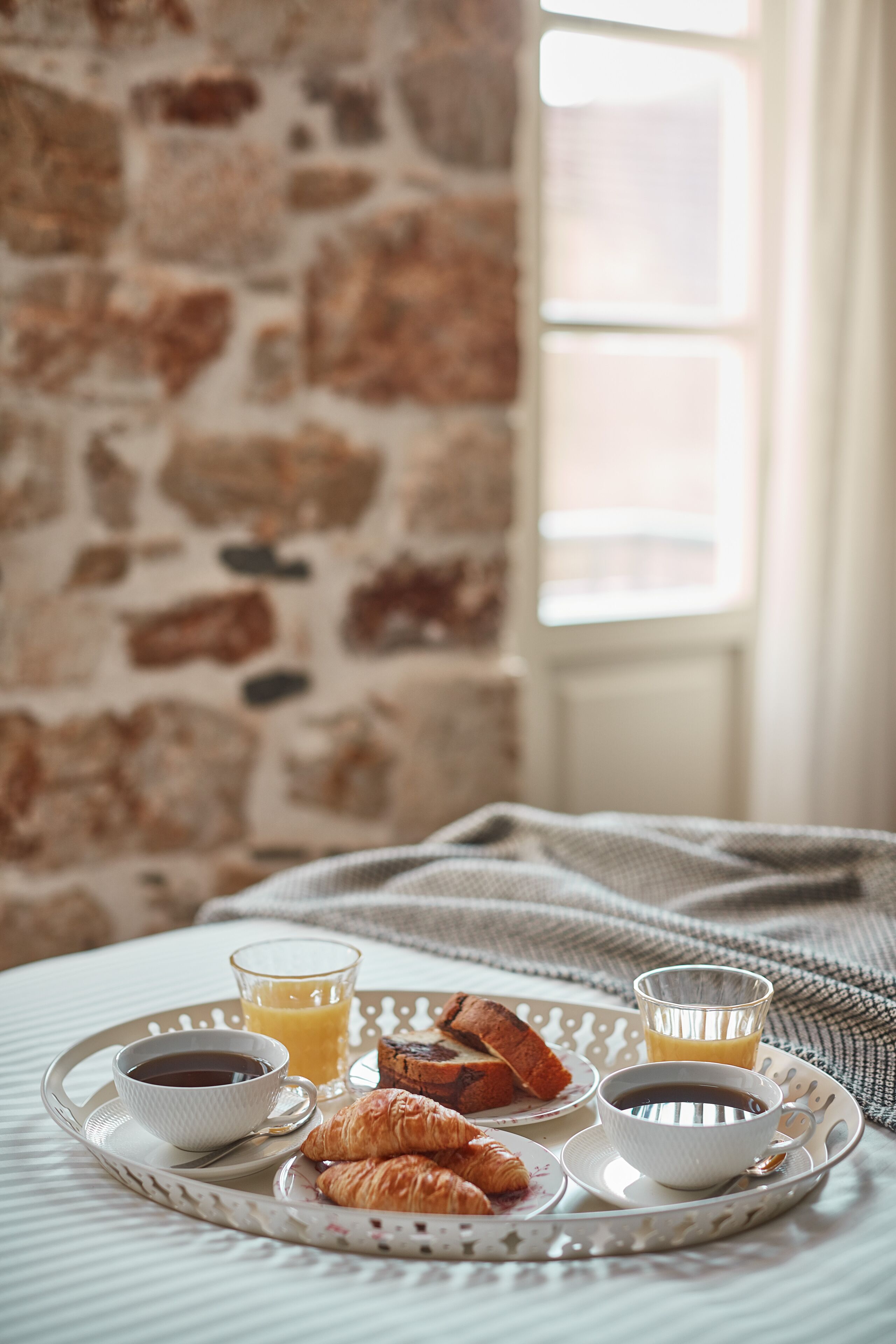 En bakke med morgenmad, blandt andet croissanter, kaffe og appelsinjuice, på en seng med en stenvæg og et vindue i baggrunden.