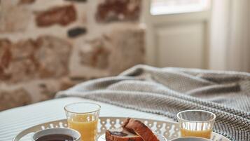 En bakke med morgenmad, blandt andet croissanter, kaffe og appelsinjuice, på en seng med en stenvæg og et vindue i baggrunden.