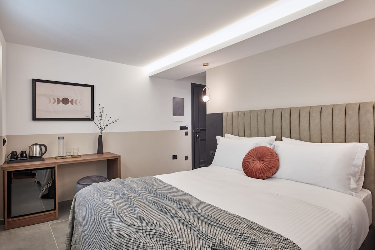 Une chambre d’hôtel moderne avec un grand lit, une table de chevet avec une lampe, un tableau encadré au mur et un petit coin salon avec un fauteuil et un vase.