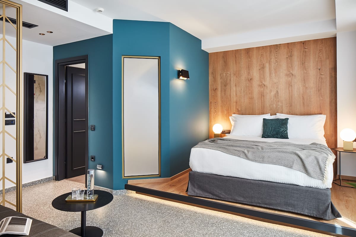 Une chambre d’hôtel moderne avec un mur d’accent de couleur turquoise, une tête de lit en bois, un grand lit avec une literie blanche et grise, et une petite table de chevet avec une lampe.