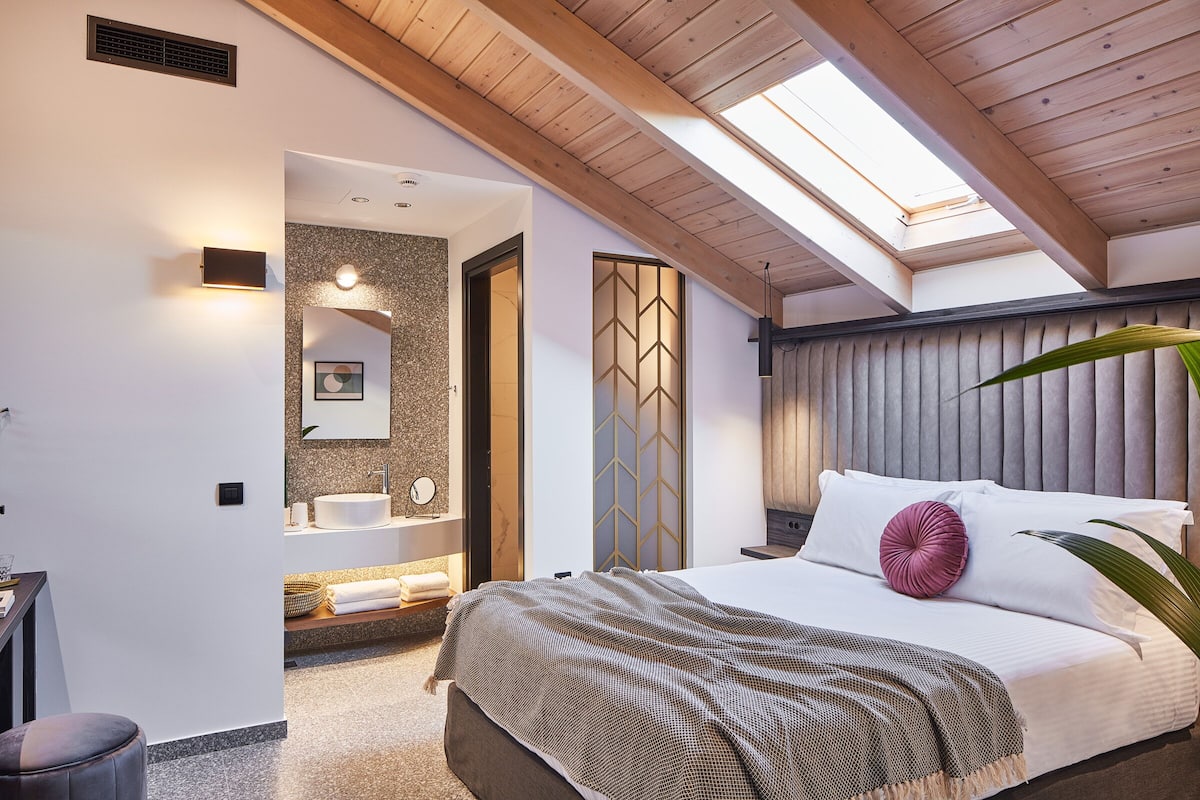 Une chambre moderne avec une lucarne, un grand lit, une table de chevet et une salle de bain équipée d’un lavabo et d’un porte-serviettes.