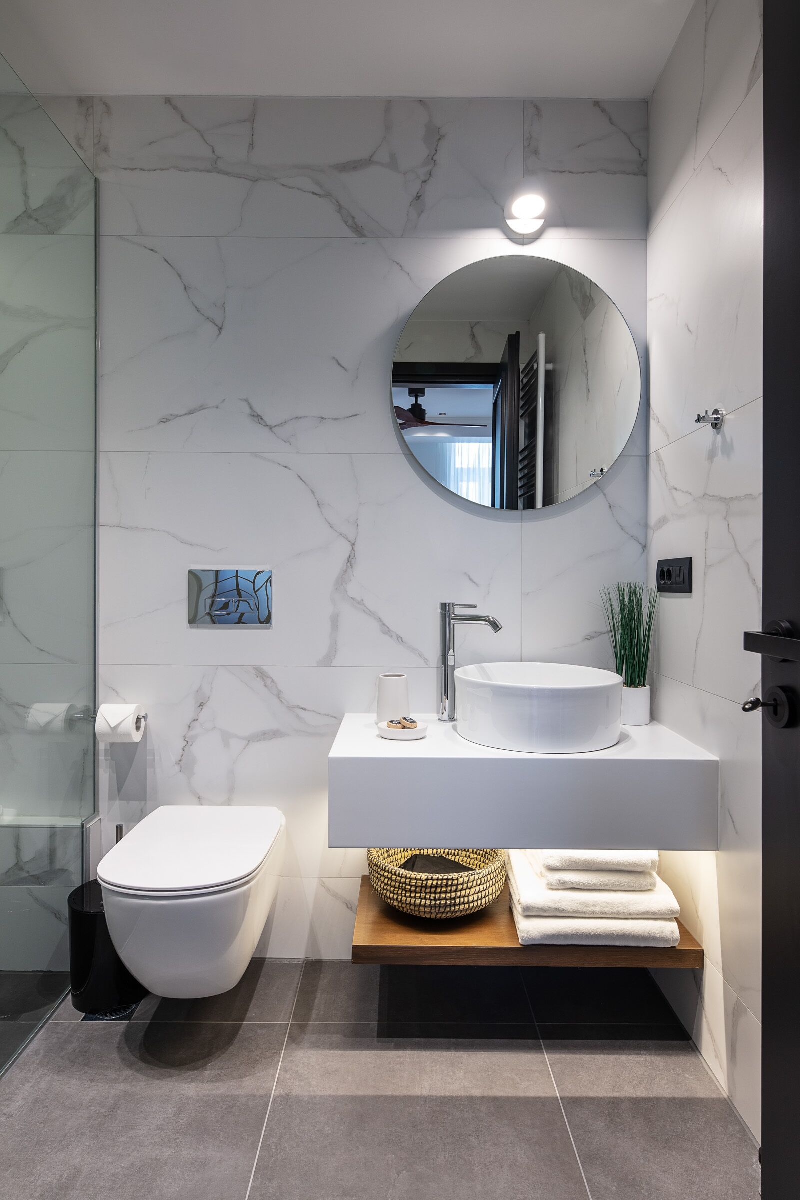 Une salle de bain moderne avec un mur en marbre, un miroir rond, un lavabo blanc et des toilettes.