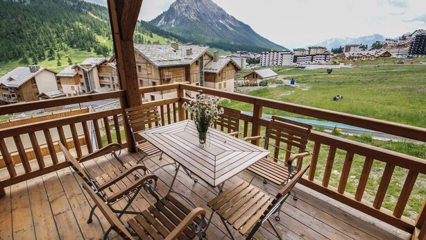 A wooden deck with a table and chairs, overlooking a mountainous landscape with buildings in the background.