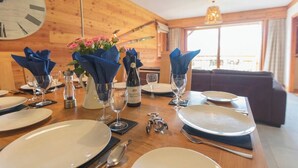 A dining table set with plates, cutlery, glasses, and a bottle of wine, with a wooden wall and a clock in the background.