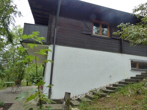 Ein zweistöckiges Gebäude mit dunkler Holzfassade und weißen Wänden, ein Fenster mit Holzrahmen sowie eine Steintreppe, die zum Eingang führt.