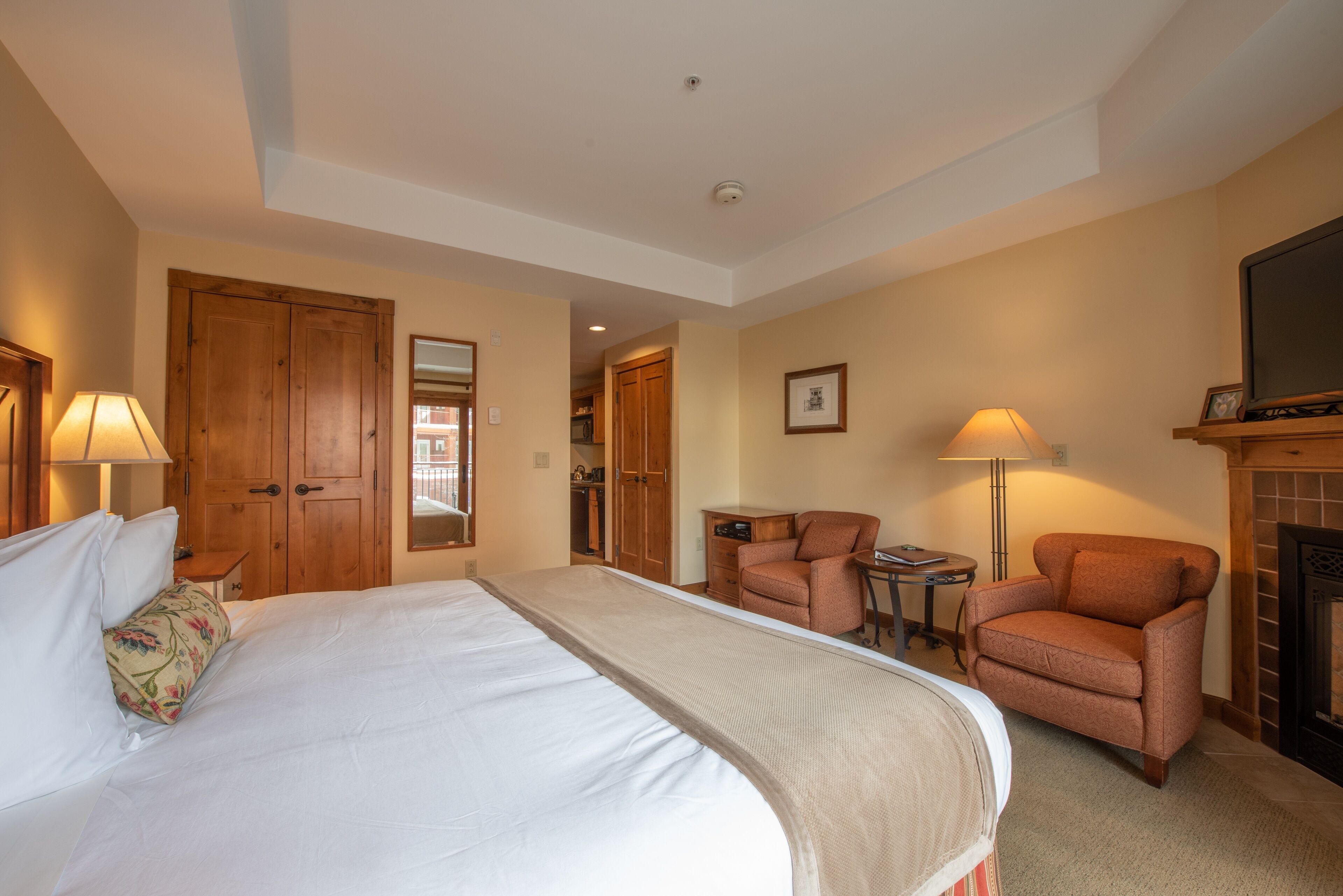 A hotel room with a large bed, two armchairs, a small table, a fireplace, and wooden doors.