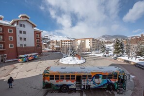 A bus stop with a colorful bus, a building with multiple floors, and a snowy mountain in the background. - Wheelchair Accessible 2-Bedroom Slopeside Condo - No Cleaning Fee! by RedAwning (Crested Butte)