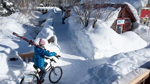A person on a bicycle with skis strapped to the back, navigating a snowy path with a wooden fence and a red building in the background. - Wheelchair Accessible 2-Bedroom Slopeside Condo - No Cleaning Fee! by RedAwning (Crested Butte)