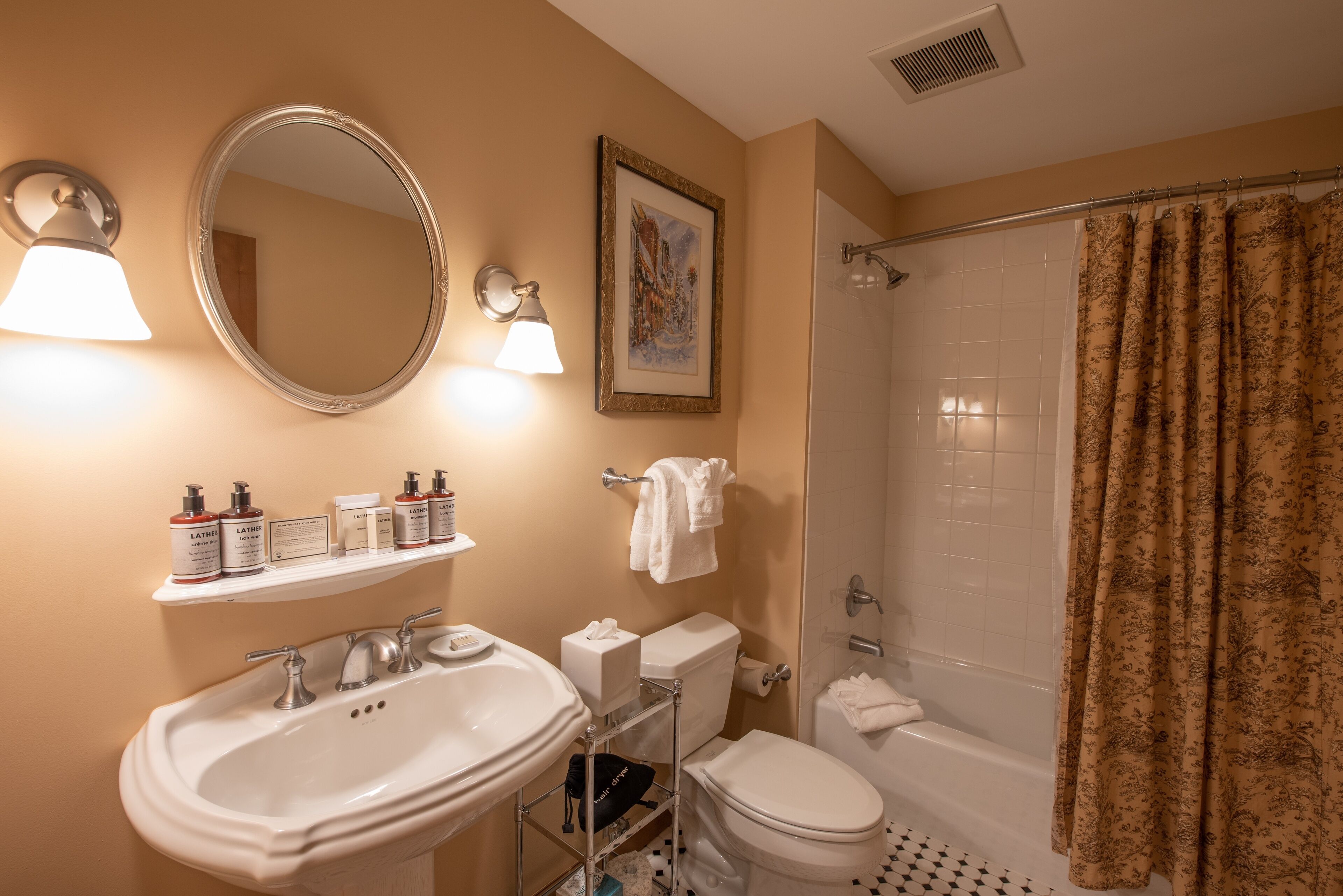 A bathroom with a sink, mirror, toilet, and shower. There are toiletries on a shelf, and a towel hangs on the wall.
