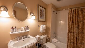 A bathroom with a sink, mirror, toilet, and shower. There are toiletries on a shelf, and a towel hangs on the wall.