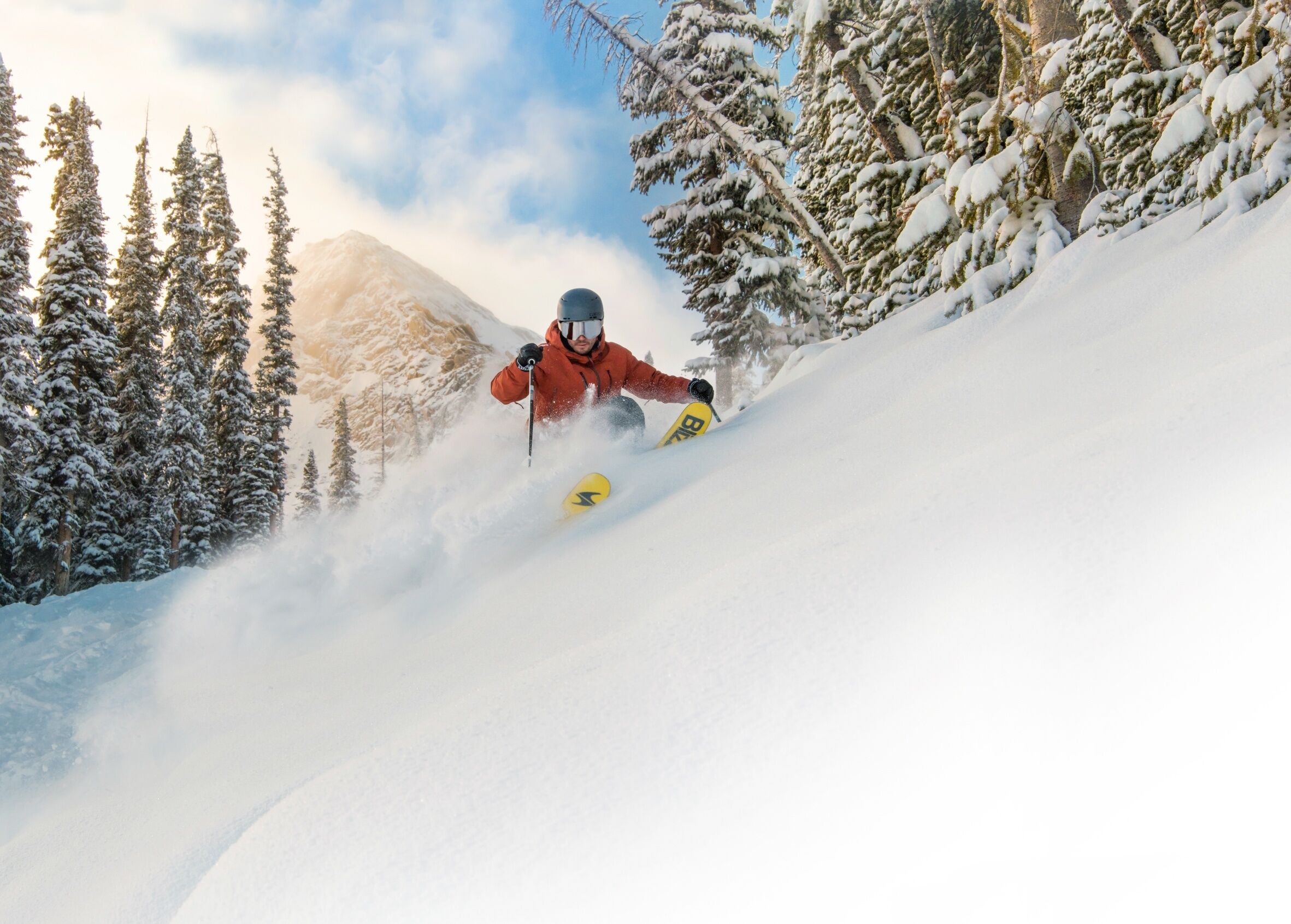 Un skieur, vêtu d’une veste rouge et d’un casque, descend une pente enneigée bordée d’arbres des deux côtés.