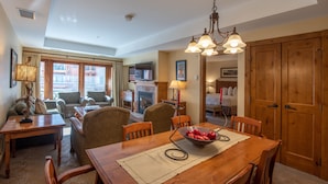A dining area with a wooden table, chairs, and a bowl of fruit. There is a fireplace, a television, and a window with curtains.