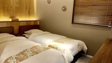 A hotel room with two single beds, wooden headboards, a wooden nightstand, a bedside table, a wall-mounted lamp, a window with blinds, and a small decorative item on the wall.