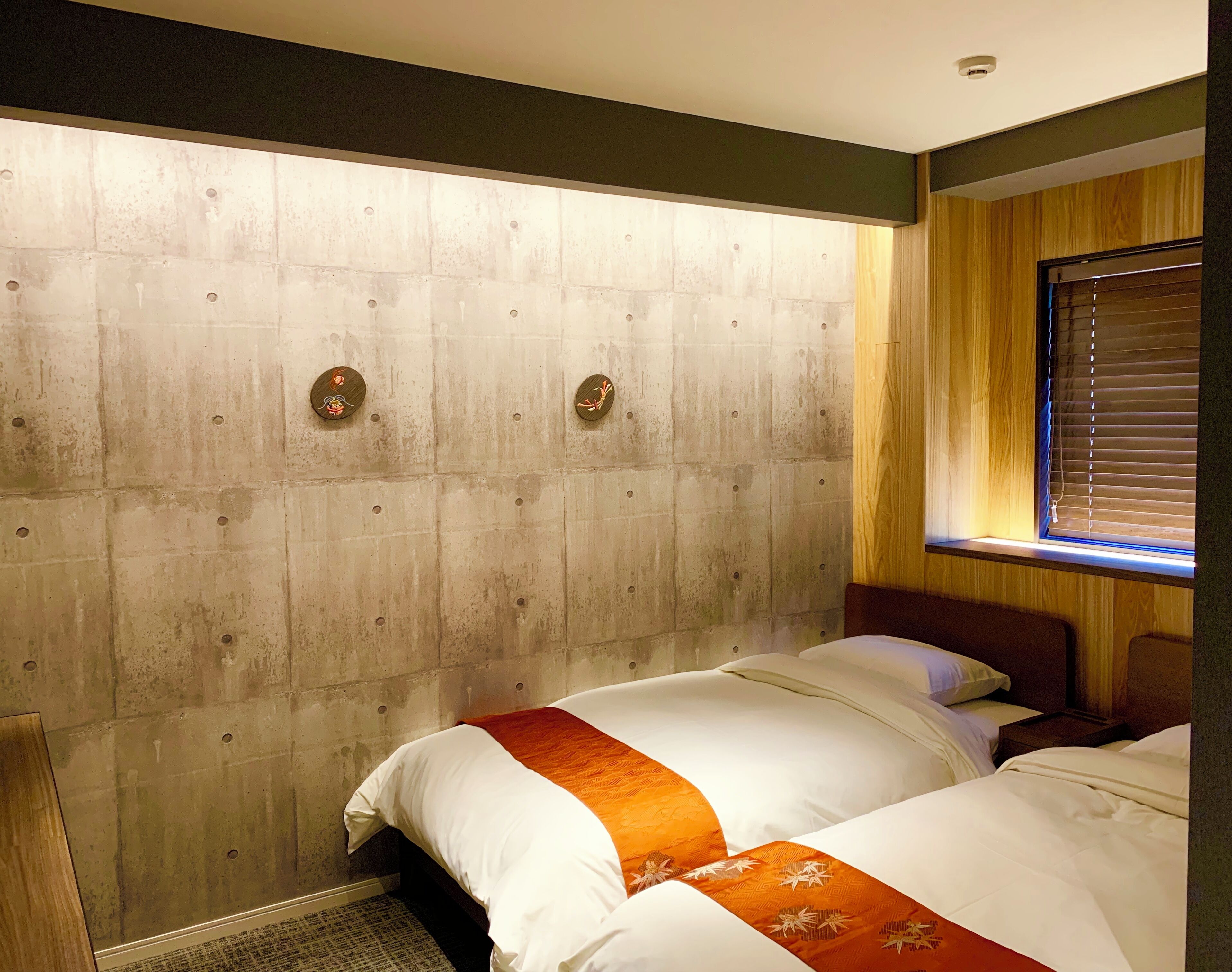 A hotel room with two beds, a wooden headboard, a window with blinds, and a visible ceiling light.