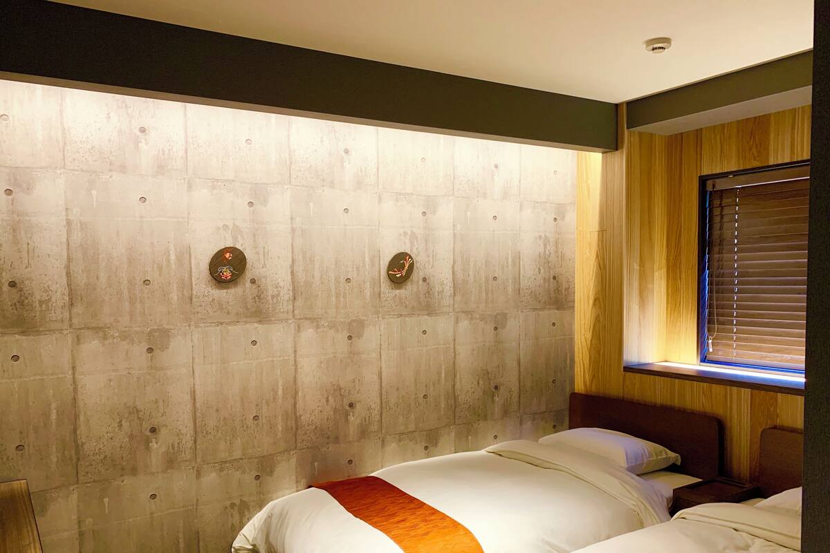 A hotel room with two beds, a wooden headboard, a window with blinds, and a visible ceiling light.