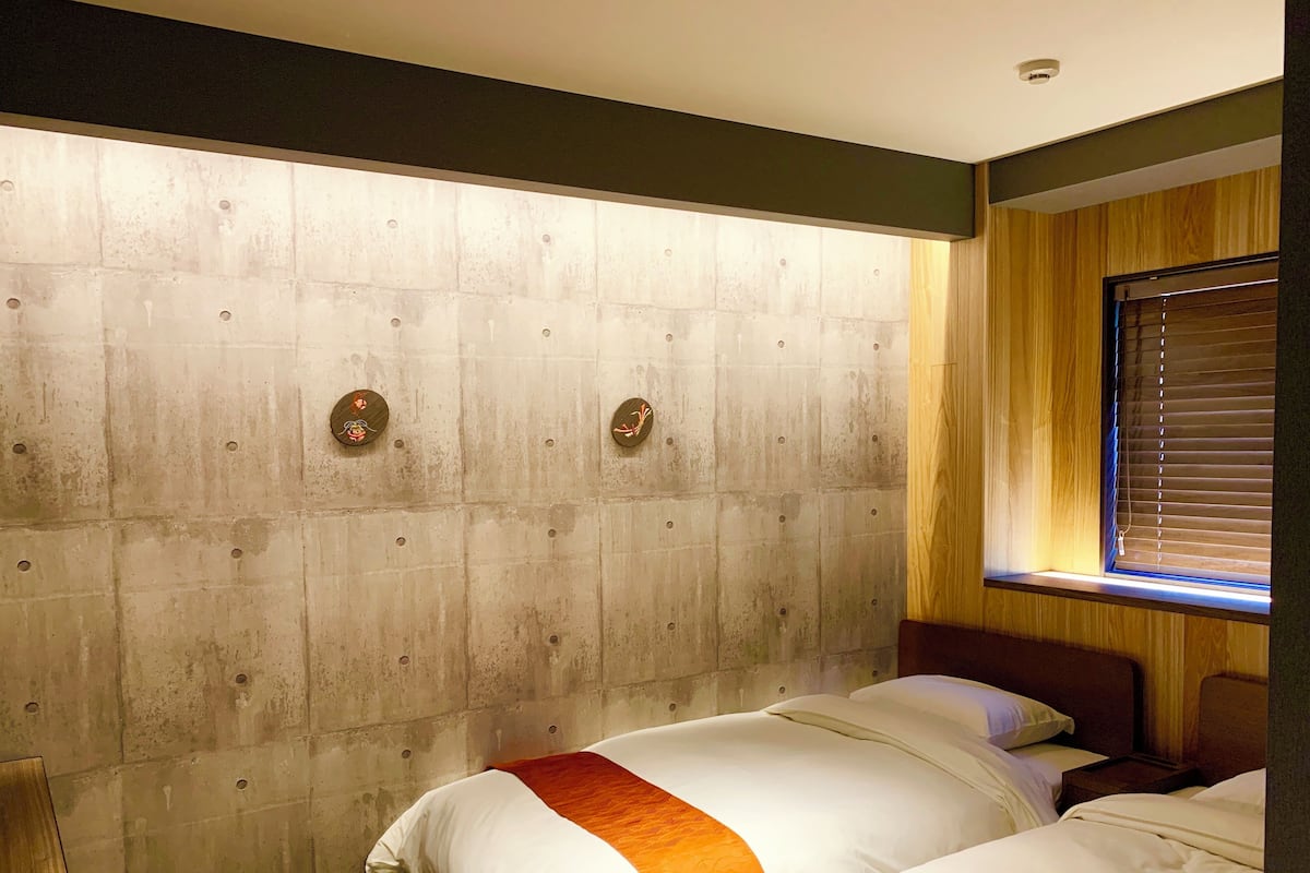 A hotel room with two beds, a wooden headboard, a window with blinds, and a visible ceiling light.