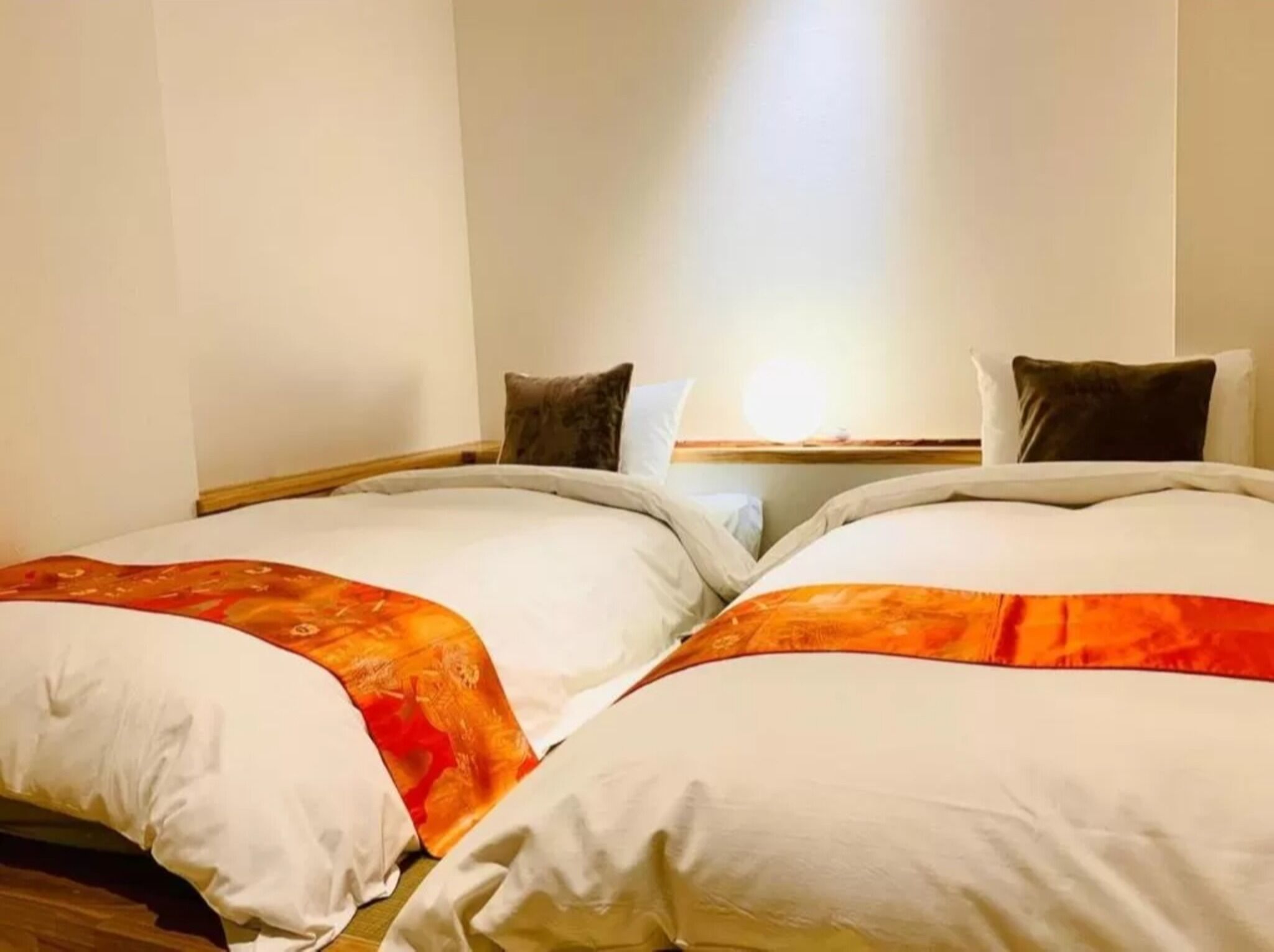 A hotel room with two beds, each with white bedding and orange accents, a wooden headboard, and a light source from a window above.