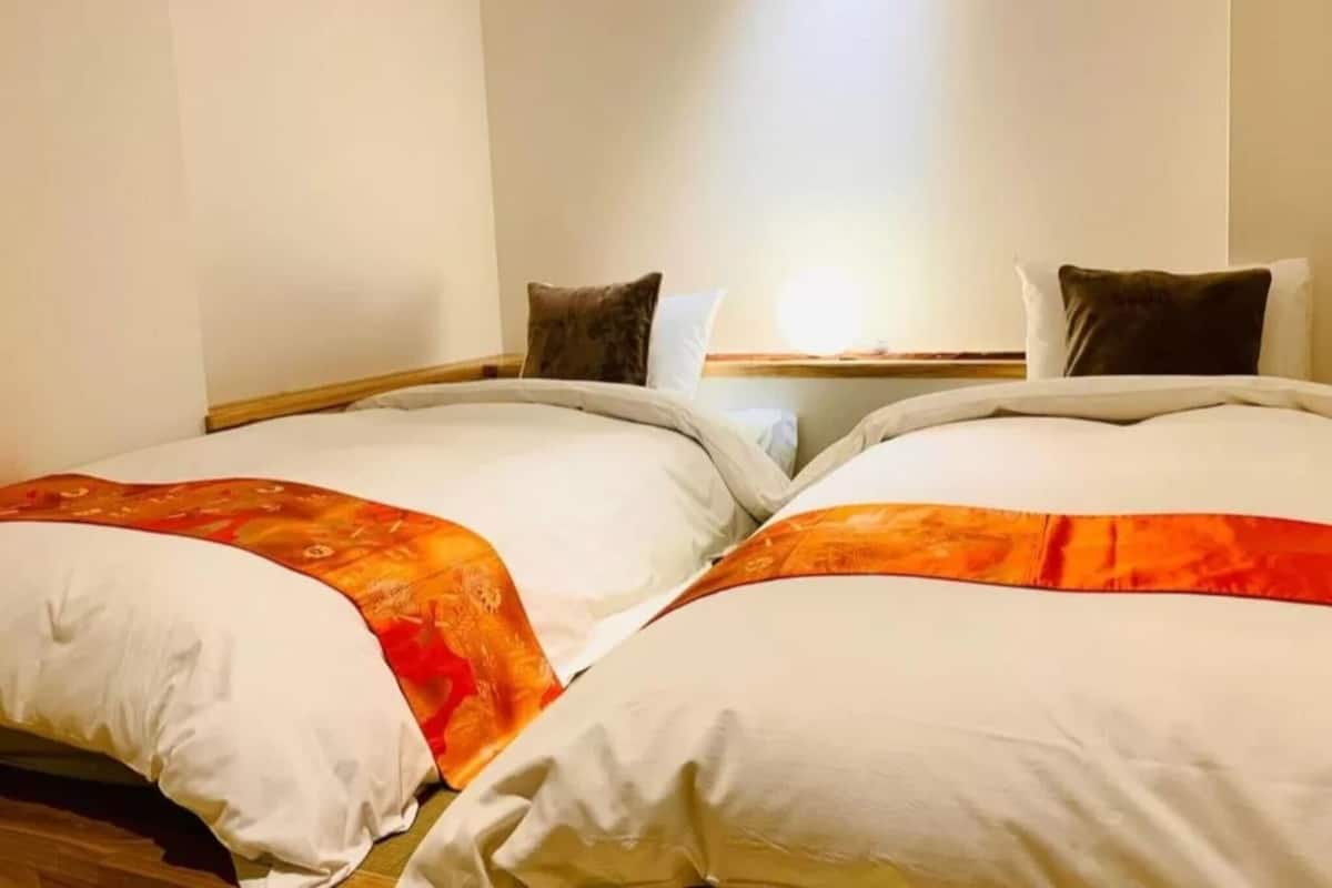 A hotel room with two beds, each with white bedding and orange accents, a wooden headboard, and a light source from a window above.