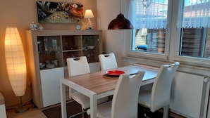 Un comedor con una mesa de madera y sillas blancas, un reloj y una lámpara decorativa sobre una estantería.